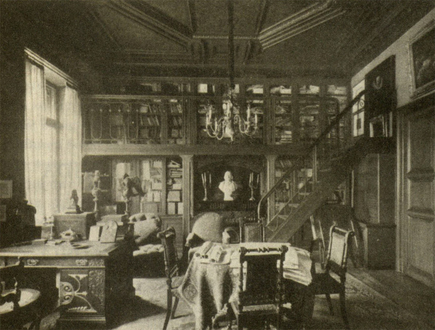 Ein Foto der Bibliothek von Johannes Fastenrath. Der Raum enthält ein großes Regal gefüllt mit Literatur, einen schreibtisch, einen weiteren Tisch und eine Treppe die zu einer weiteren Etage führt.