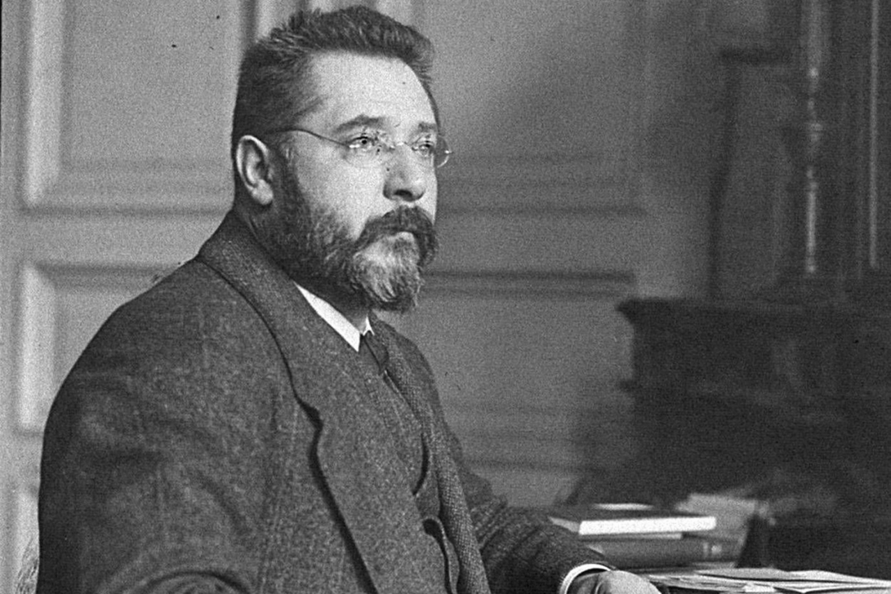 Ein Porträt von Heinrich Erkes. Er ist angelehnt an einem Schreibtisch, mit dem Blick zur Seite. Er trägt einen Anzug und eine Brille.