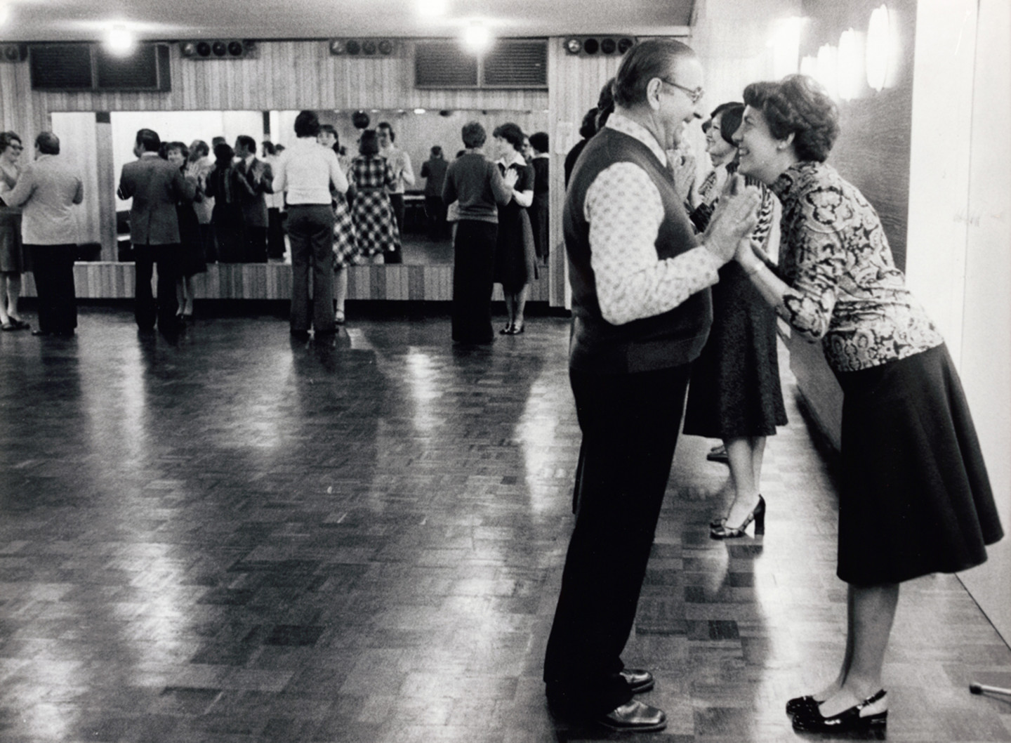 Foto von Menschen in einer Tanzschule. Im Fokus stehen eine Frau und ein Mann, die sich gegenüber stehen und sich anlächeln.