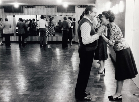 Photo of people in a dance school. The focus is on a woman and a man standing opposite each other and smiling at each other.