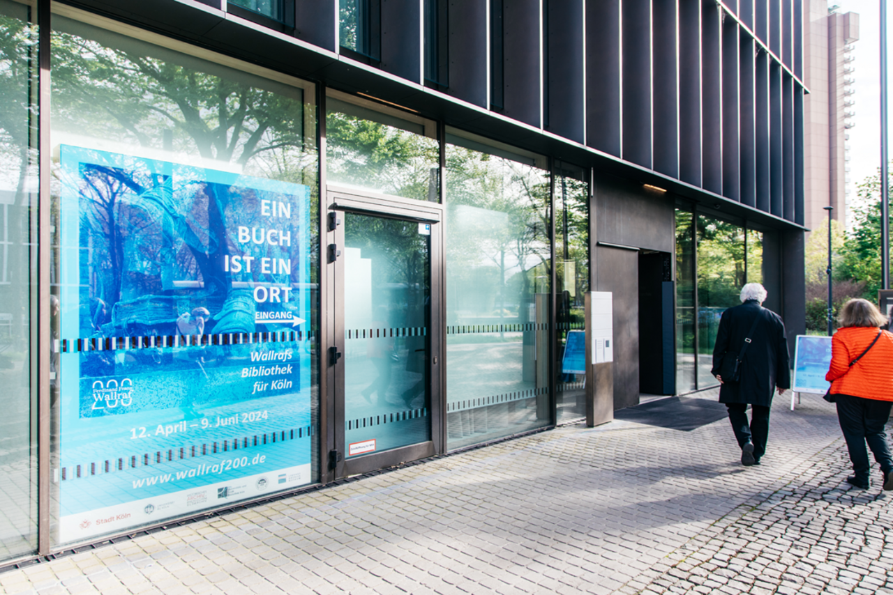 Besucher*innen der Ausstellungseröffnung gehen auf den Eingang des Stadtarchivs zu. Im Vordergrund ist ein großes Ausstellungsplakat in der Fensterfront des Stadtarchivs zu sehen.