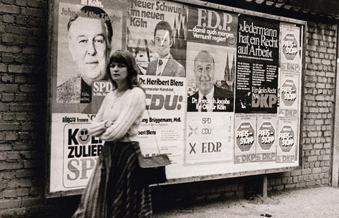 A woman walks while looking into the camera. In the background are advertising posters of politicians for the 1975 state and local elections.