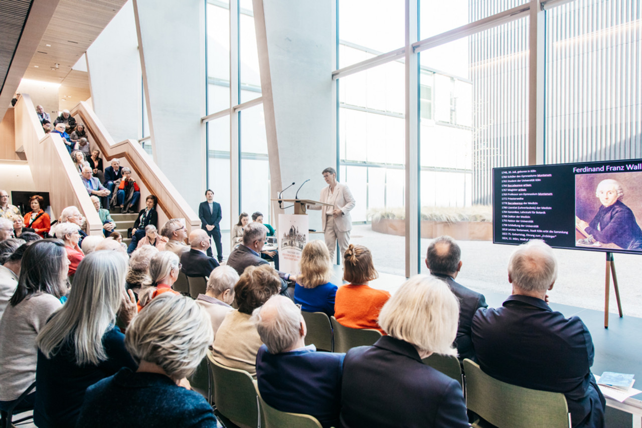 Das Foyer des Stadtarchivs ist gefüllt mit Besucher*innen der Ausstellungseröffnung, die der Rednerin Dr. Christiane Hoffrath zuhören.