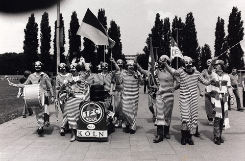 Photo of costumed fans of 1.FC Köln, on their way to the stadium. They are dressed like clowns, wear striped clothes and hold flags and drums in their hands.