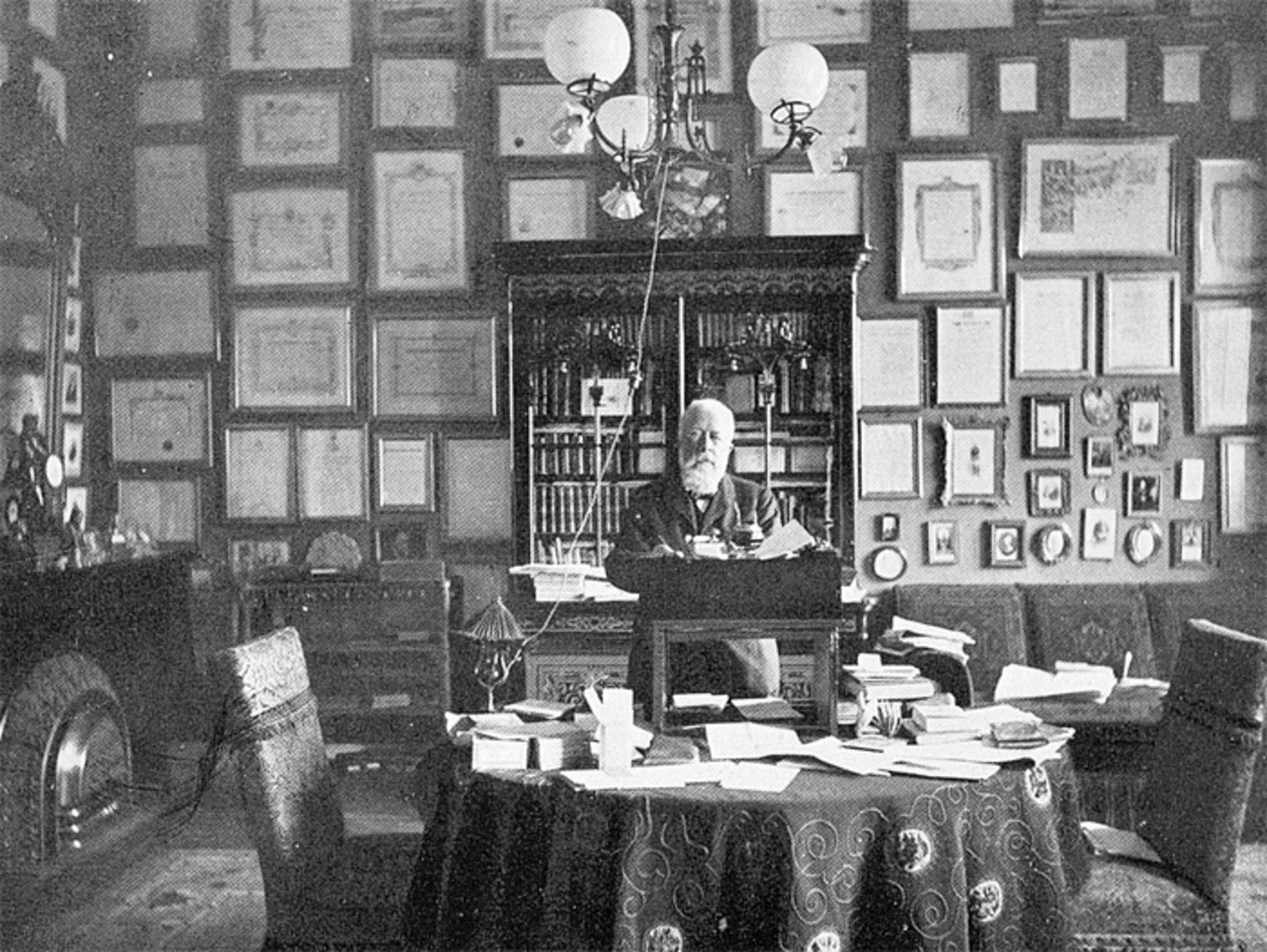 Ein Foto von Johannes Fastenrath in seinem Arbeitszimmer wie er an einem Schreibtisch sitzt, den Blick zur Kamera gerichtet. Die Wand hinter ihm ist gefüllt mit Bilderrahmen und an der Seite ist ein Kamin zu erkennen.