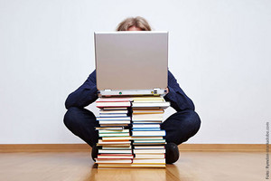 Man sieht einen Bücherstapel auf dem ein Laptop steht, dahinter sitzt eine Person.