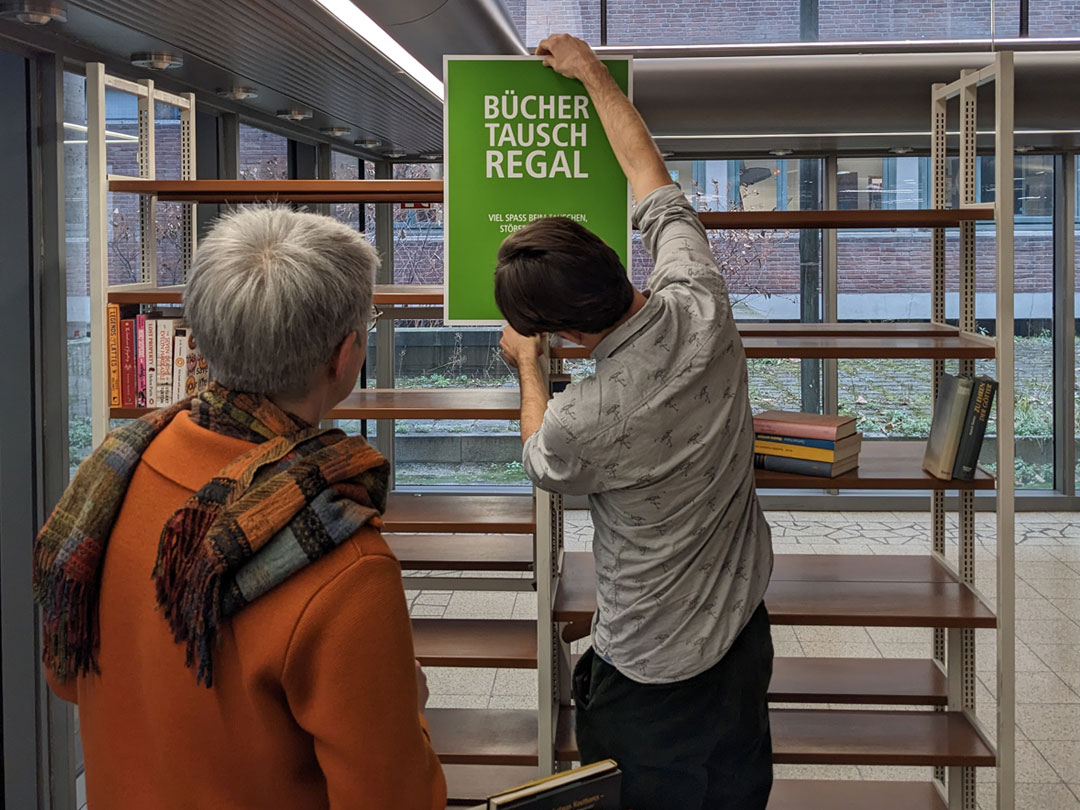 A photo with two people preparing the book exchange shelf in the foyer of the UCL Cologne.