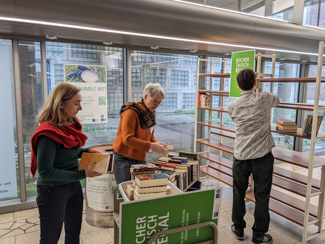 A photo of three people filling the book exchange shelf in the foyer of the UCL Cologne.
